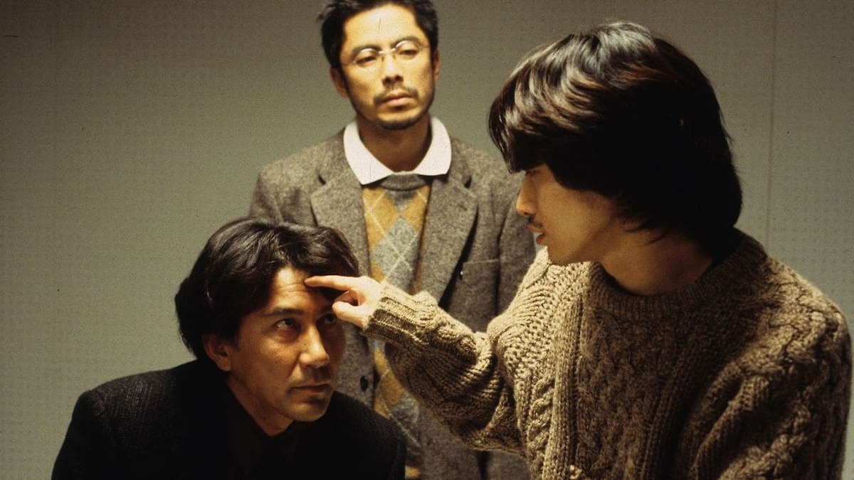 Three men in a plain-walled room: a seated man in a dark jacket looks up while a younger man in a cable-knit sweater points a finger at his forehead, and a bespectacled man in a tweed blazer and argyle sweater stands watching in the background.