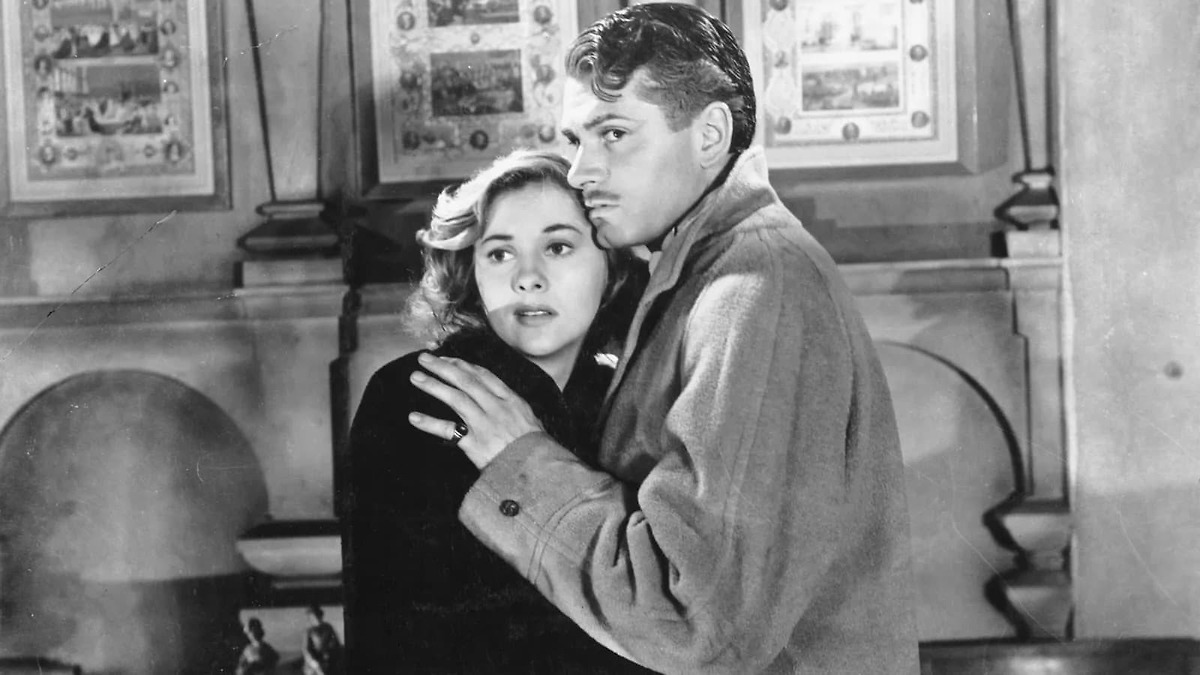 A vintage black-and-white film still shows a young blonde woman (Mrs. Winter) held protectively in the arms of a dark-haired man in a heavy wool coat (Mr. Winter); both gaze anxiously off to the side, standing in a room with framed pictures and wood paneling behind them.