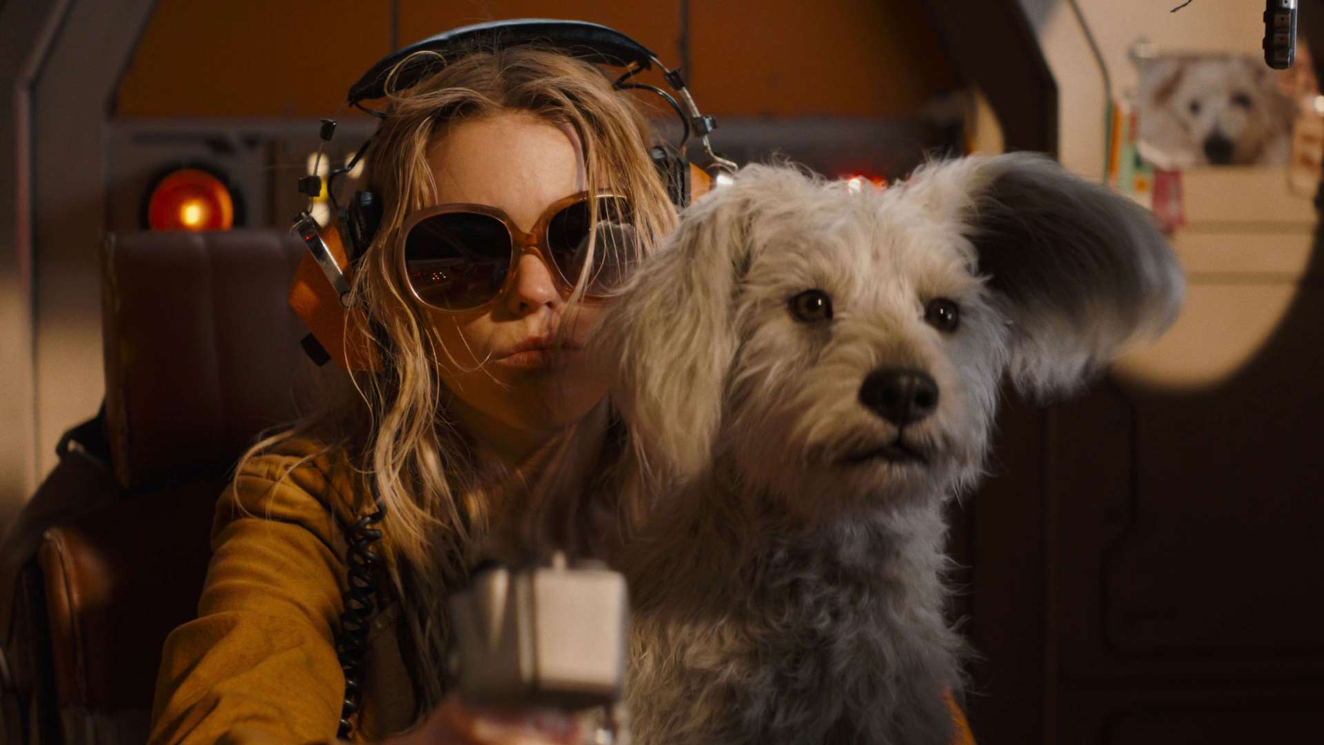 Super Girl wearing large tinted sunglasses and aviation headphones leans forward in a dimly lit cockpit-like cabin, one hand on a control lever, beside a shaggy white terrier-type dog (Krypto) with one ear flopped back, with a photo of a similar dog pinned to the wall behind them.
